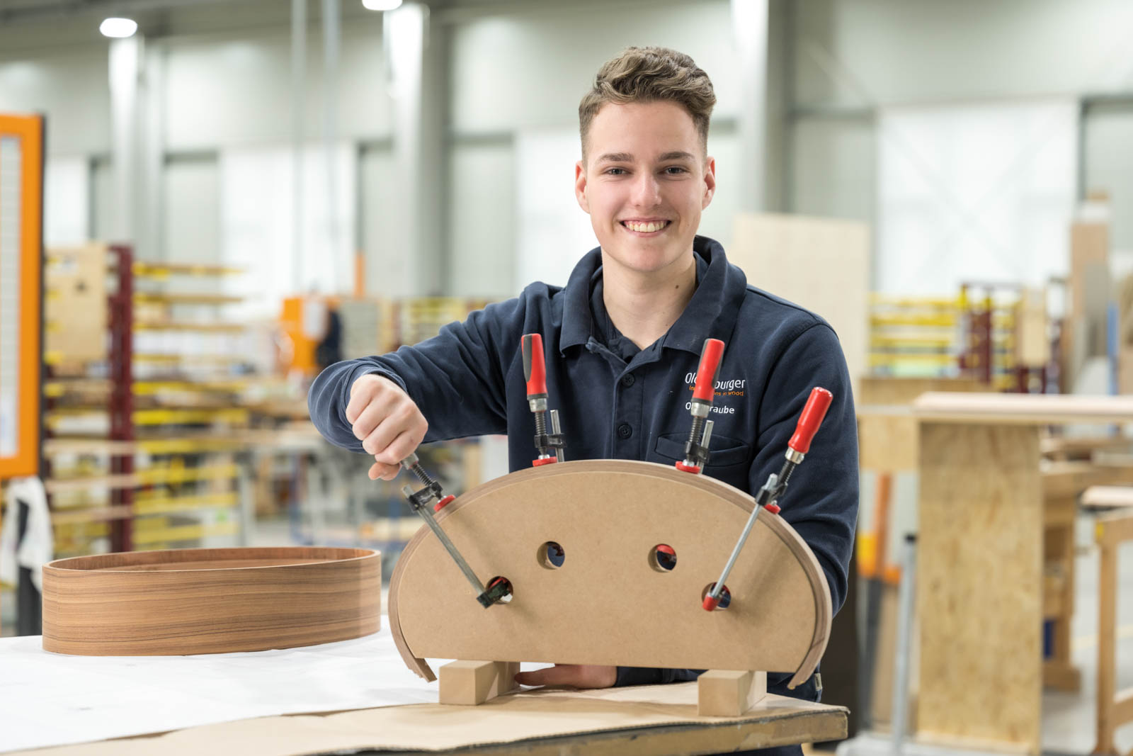 Junger Tischler in der Ausbildung spannt ein Formteil mit roten Zwingen und lächelt in die Kamera
