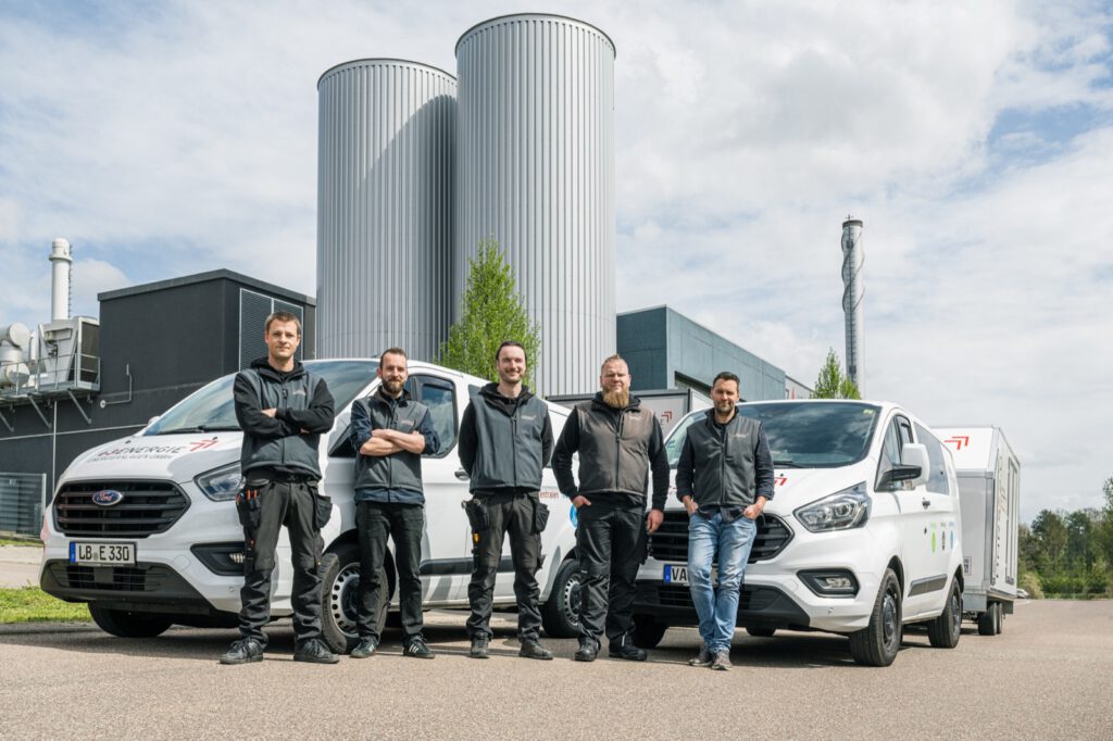 Fünf Personen von e3 vor den Servicefahrzeugen und dem Anhänger, dahinter das Heizwerk