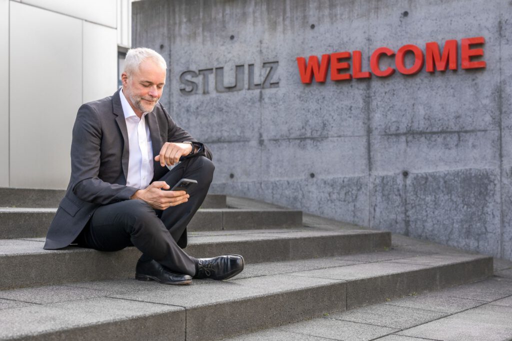 Mann sitzt auf den Stufen vor der Eingangswand von STULZ und schaut auf sein Smartphone