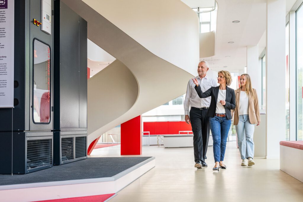 Drei Personen gehen durch das helle Foyer von STULZ mit der geschwungenen Treppe