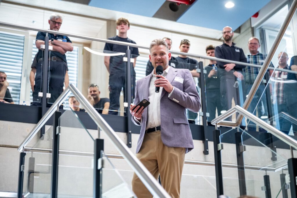 Redner mit Mikrofon auf einer Treppe, auf der Galerie darüber hören Mitarbeitende zu