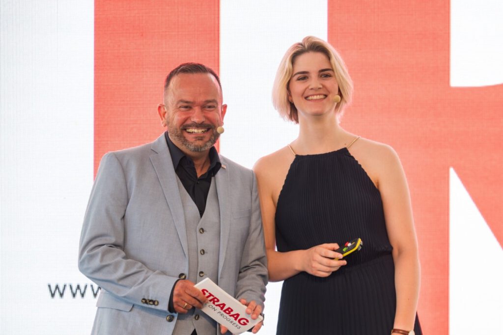 Melanie Hegemann und Andreas Thull, die Moderation der Eröffnung, lachend vor der STRABAG-Leinwand