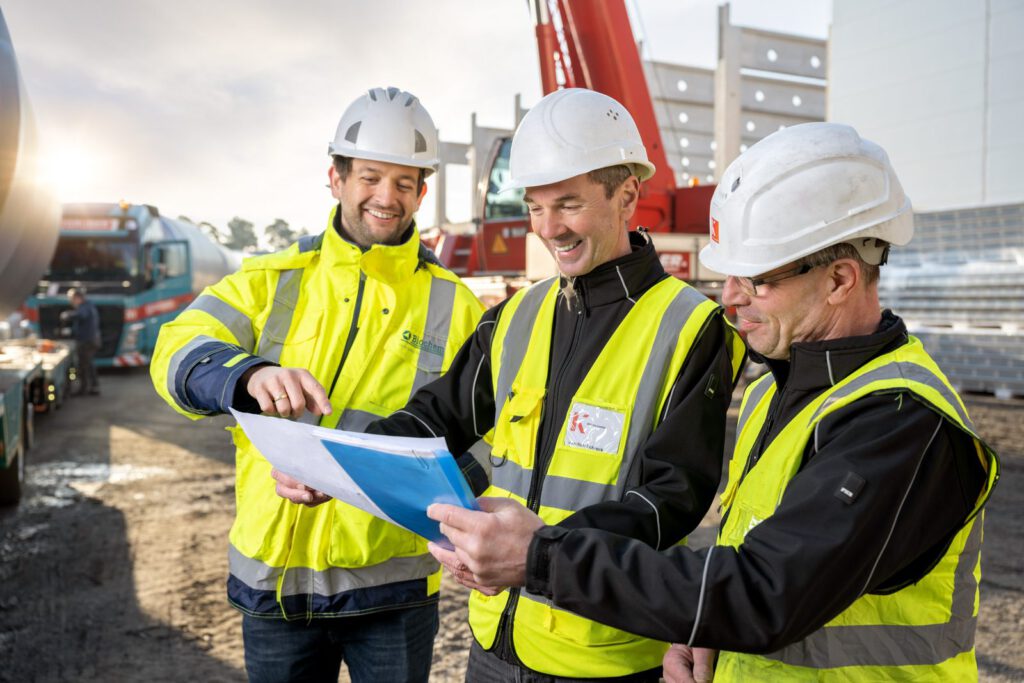 Drei Männer mit Helm und Warnweste besprechen einen Plan, dahinter Autokran und Tieflader