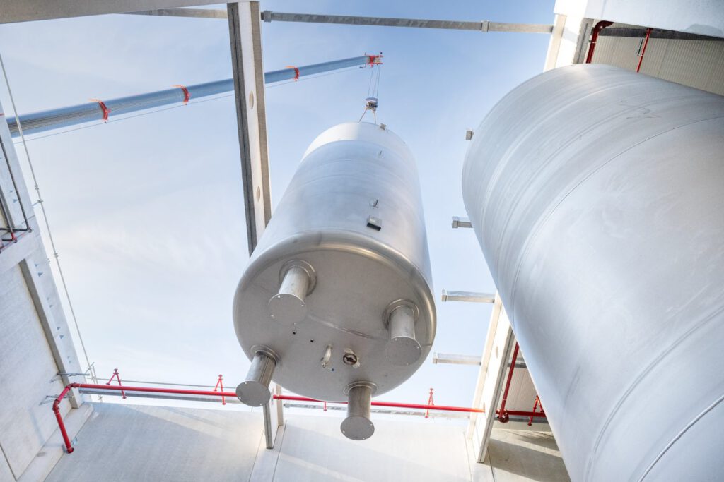 Edelstahltank schwebt am Kran durch eine Öffnung im Dach in eine neue Halle, Blick von innen nach oben