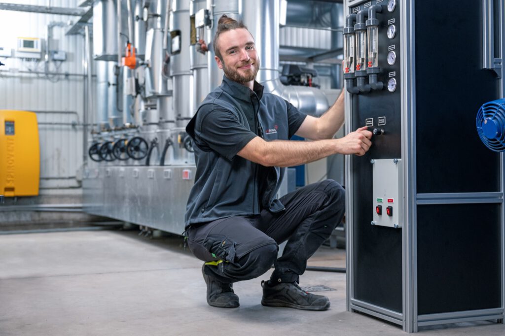 Techniker von e3 an der Messstation einer Heizzentrale, dahinter Pumpen und Rohrleitungen