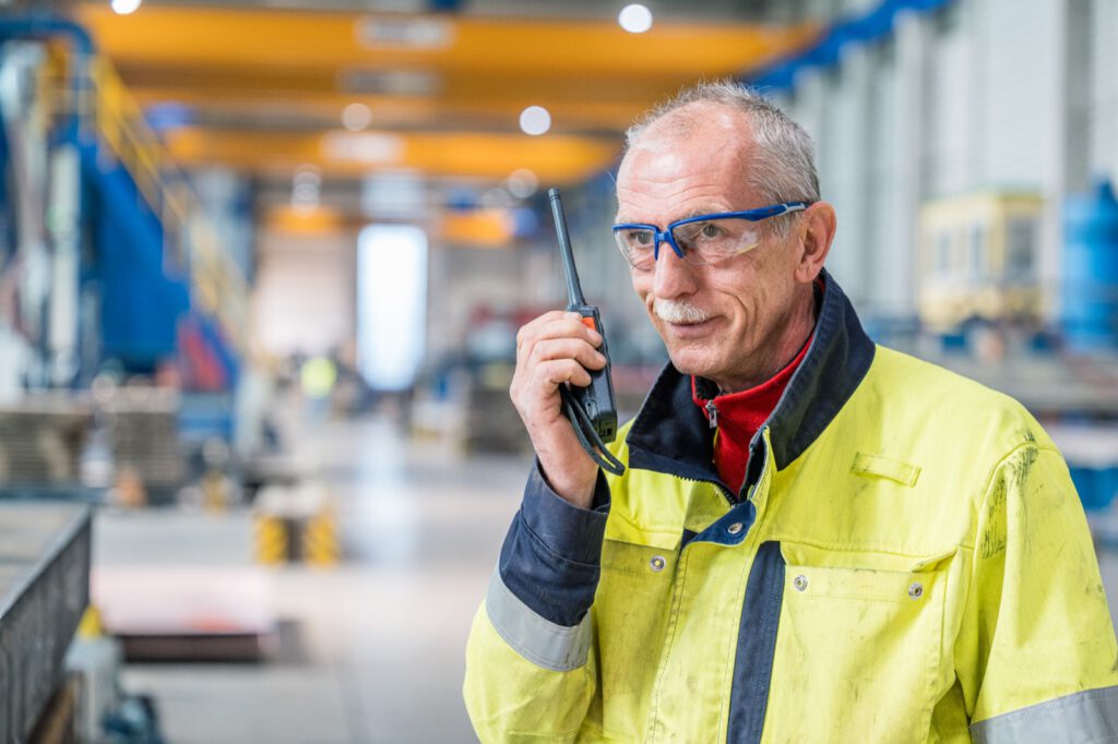 Mitarbeiter mit Schutzbrille und Warnjacke spricht in der Halle in ein Funkgerät