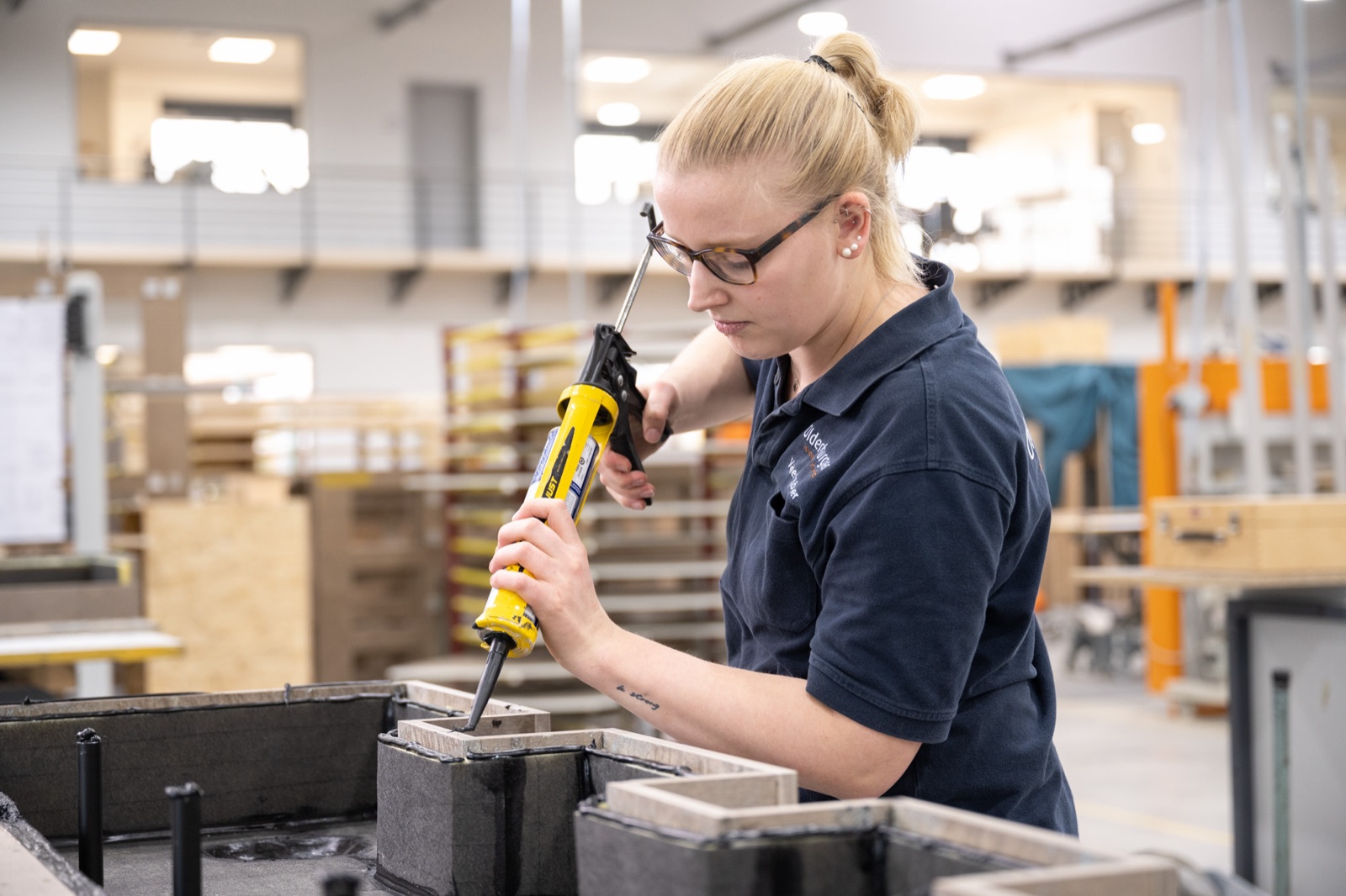 Mitarbeiterin mit Brille arbeitet konzentriert mit einer Heißklebepistole an einem Bauteil