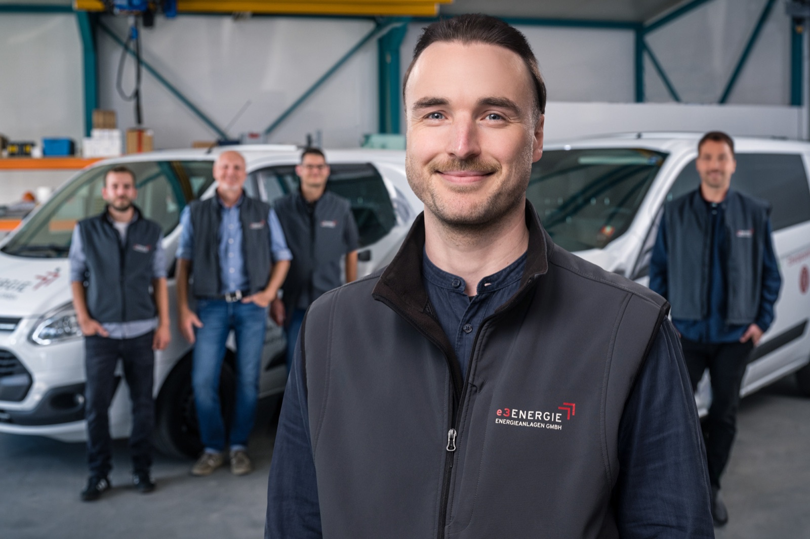 Portrait eines Mitarbeiters von e3 in Arbeitsjacke mit Logo, dahinter vier Kolleginnen und Kollegen vor den Servicefahrzeugen