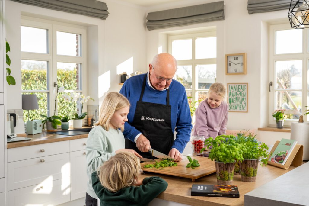 Großvater schneidet mit zwei Kindern in der Küche Kräuter, daneben Kräuter in Töpfen