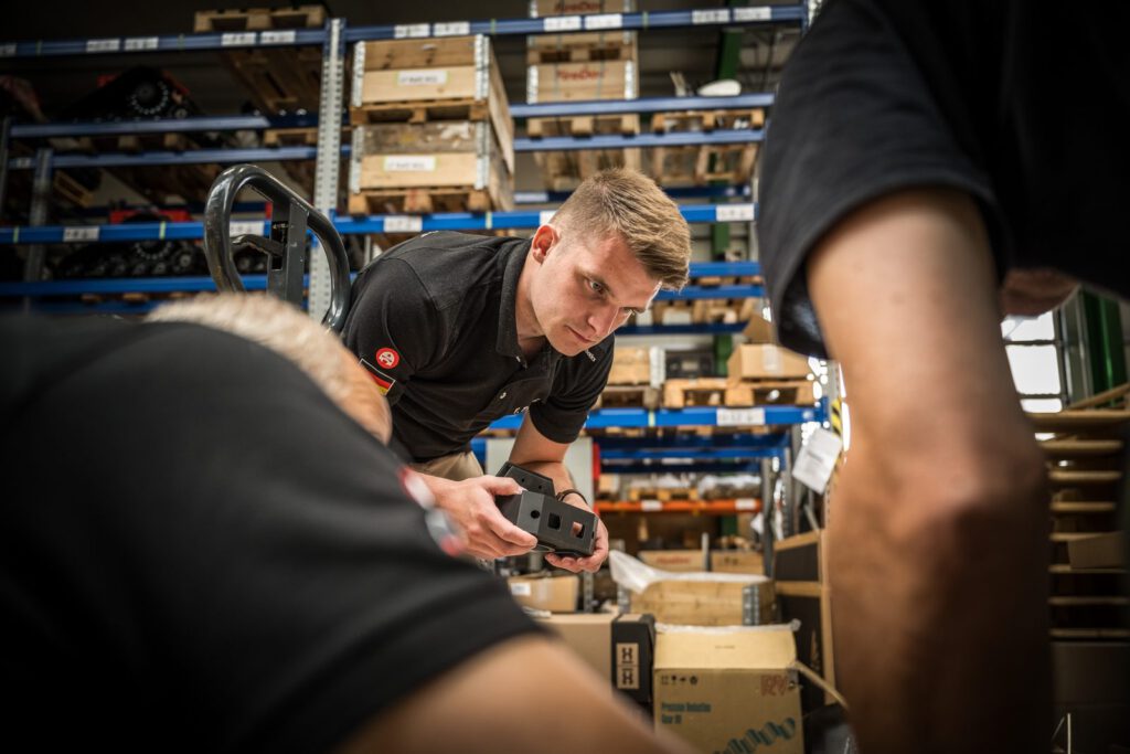 Ein Mitarbeiter von Alpha Robotics mit einem Bauteil in der Halle, im Hintergrund Regale