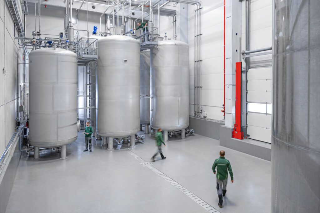 Drei große Edelstahltanks im Tanklager von Biochem, Mitarbeitende in grünen Jacken gehen durch die Halle