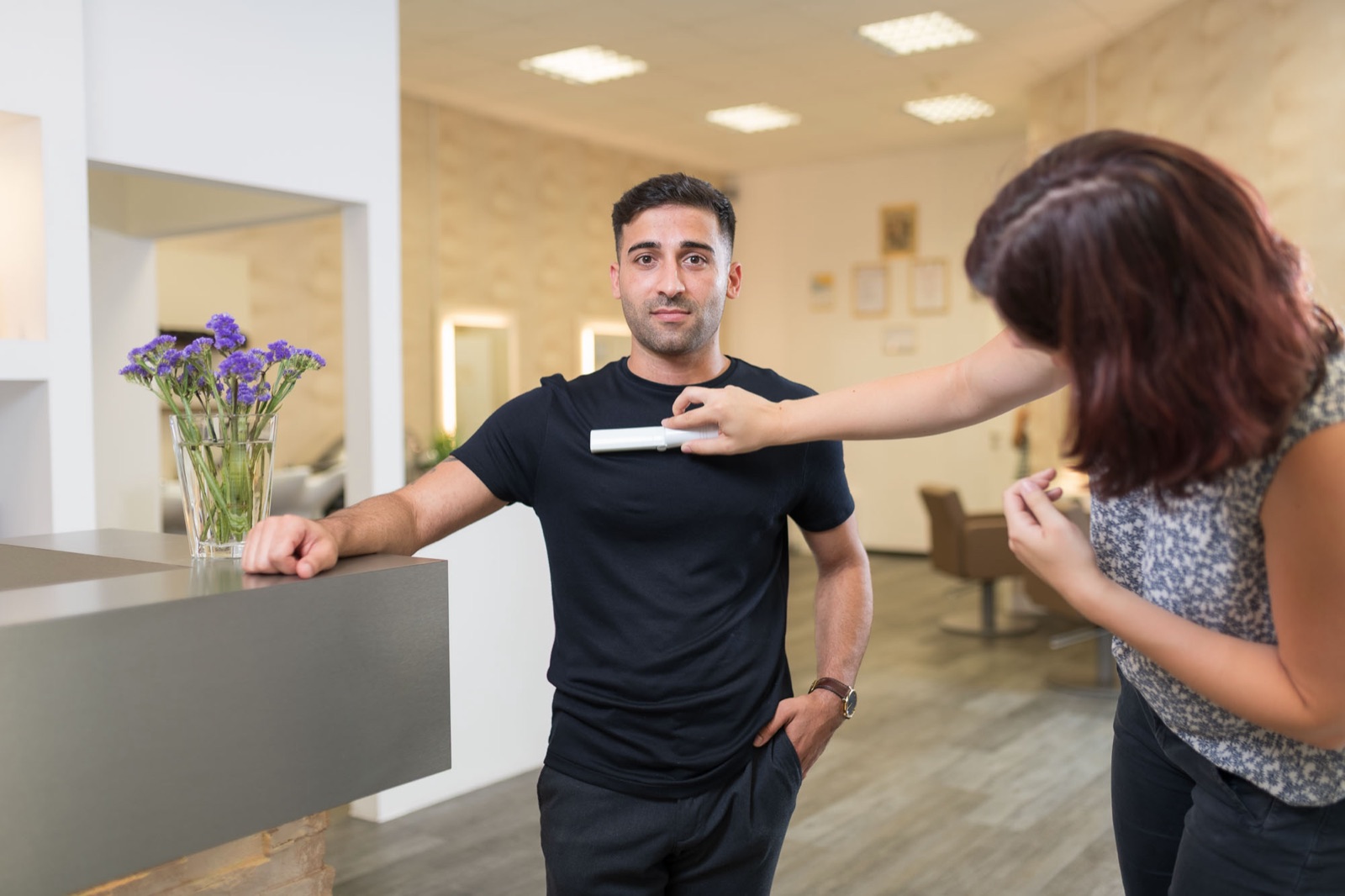 Astrid entfernt mit einer Fusselrolle Flusen vom schwarzen Shirt eines Mannes, der im Friseursalon an einer Theke lehnt