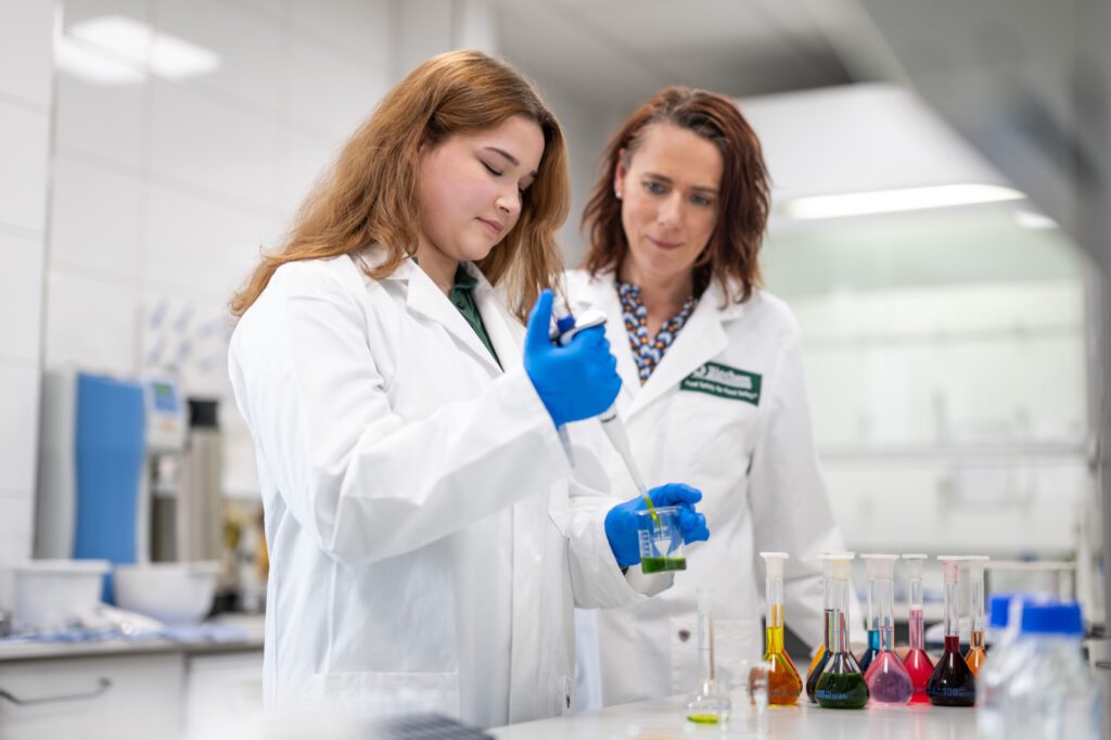 Eine Auszubildende im Laborkittel arbeitet mit Pipette und Handschuhen, eine Kollegin schaut zu
