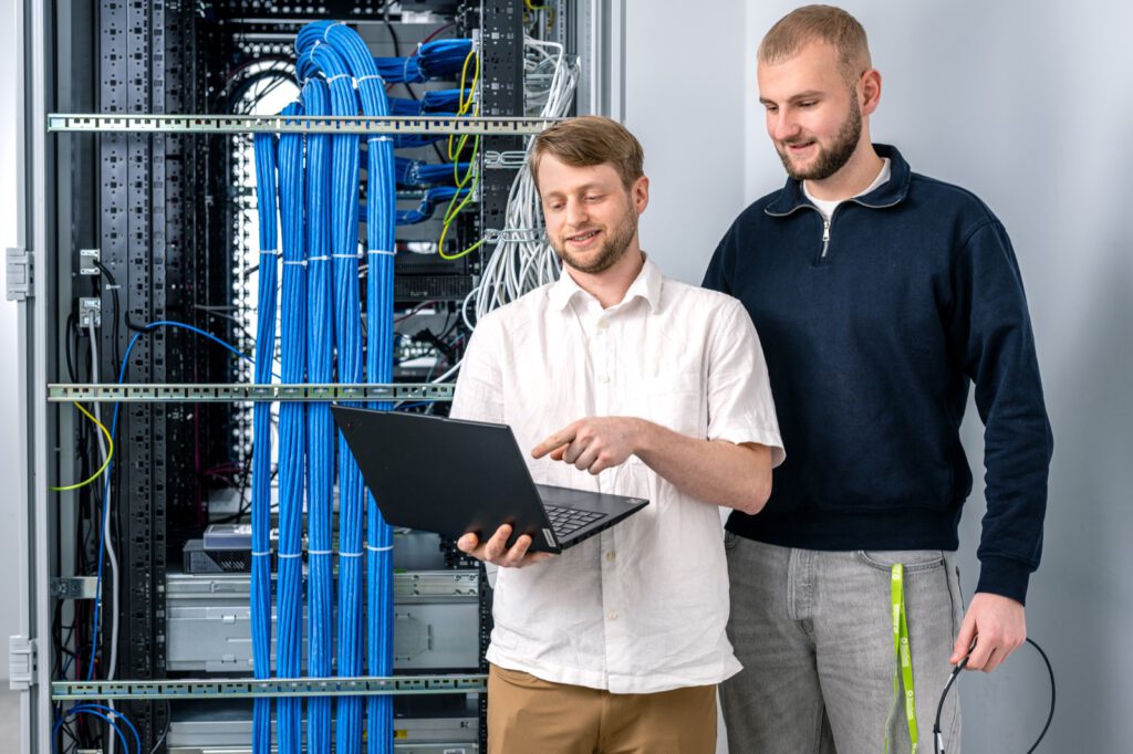 Zwei junge Männer mit Laptop vor einem Serverschrank mit blauen Kabeln