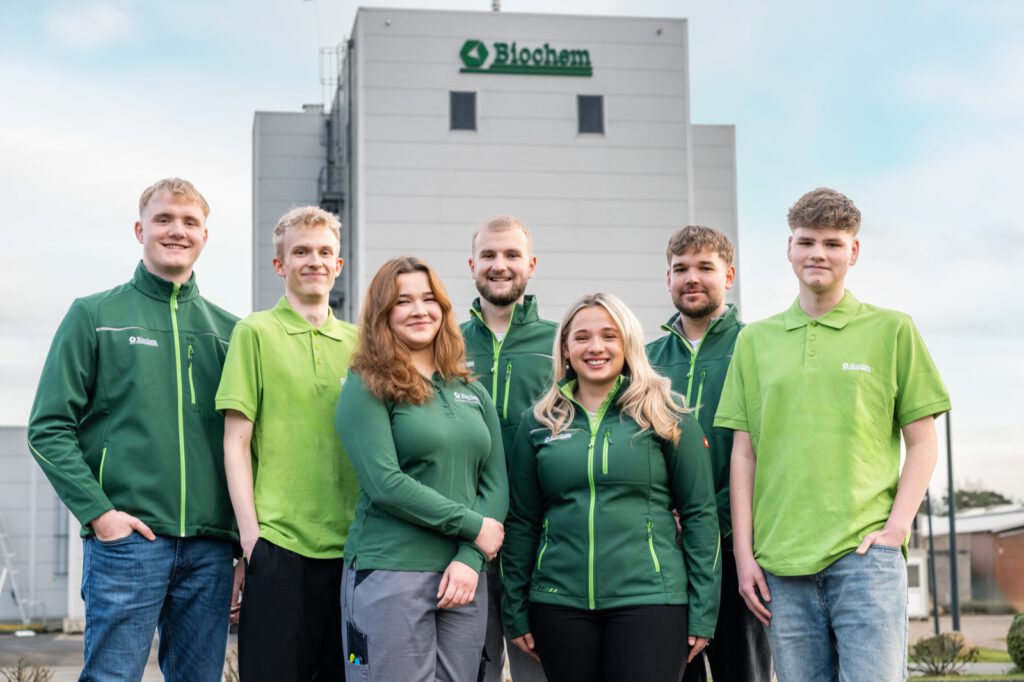 Die Auszubildenden von Biochem in grünen Jacken und Poloshirts vor dem Werksturm mit Logo
