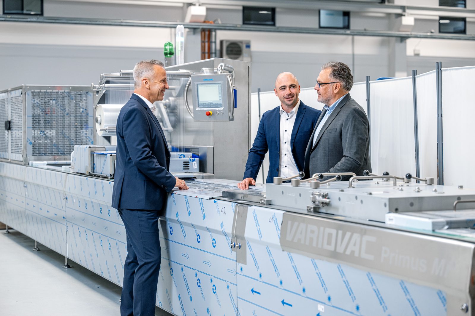 Drei Männer in Anzügen im Gespräch an einer Verpackungsmaschine in der Halle